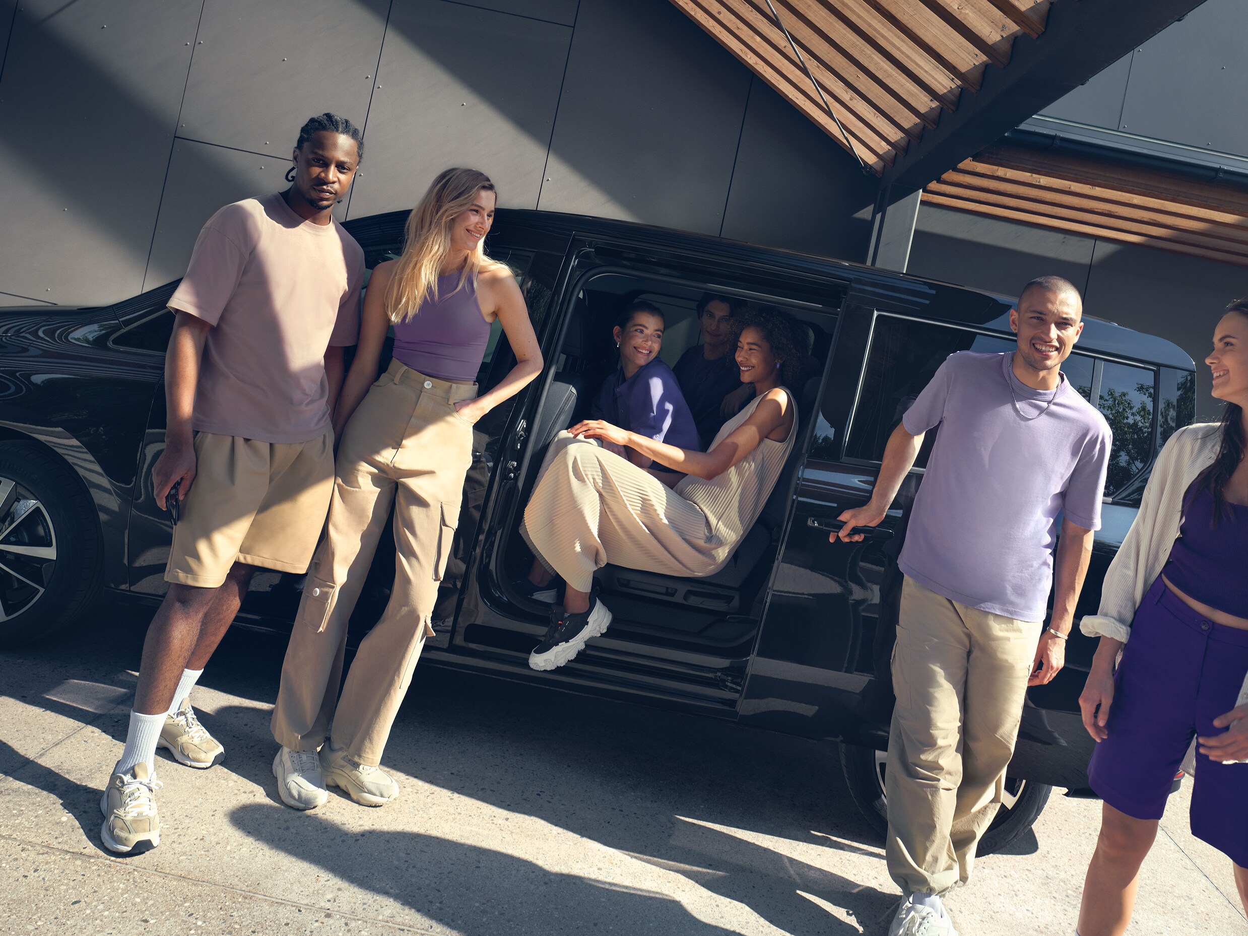 Groep jonge vrienden stapt in een zwarte Mercedes-Benz T-Klasse geparkeerd voor een modern gebouw, genietend van een zonnige dag.