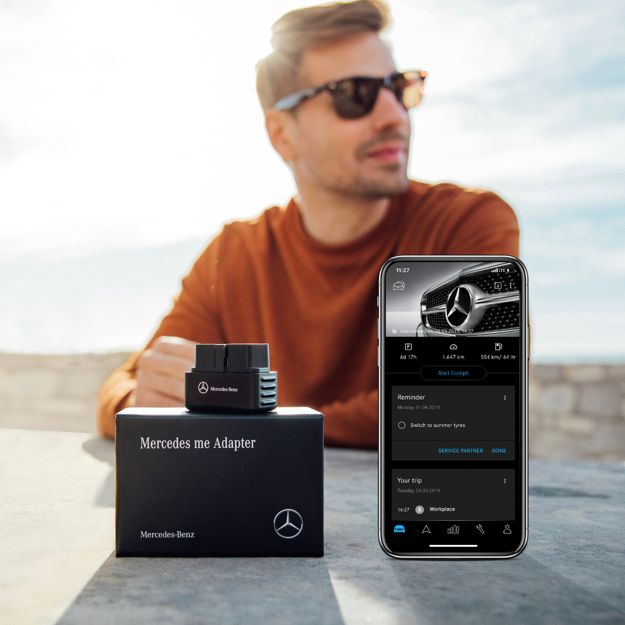 Détection d'accident avec le Mercedes-Benz Adapter. | Mercedes-Benz Un homme portant des lunettes de soleil est assis à une table sur laquelle se trouvent, au premier plan, des adaptateurs Mercedes-Benz et un smartphone avec l'application Mercedes-Benz Adapter ouverte.