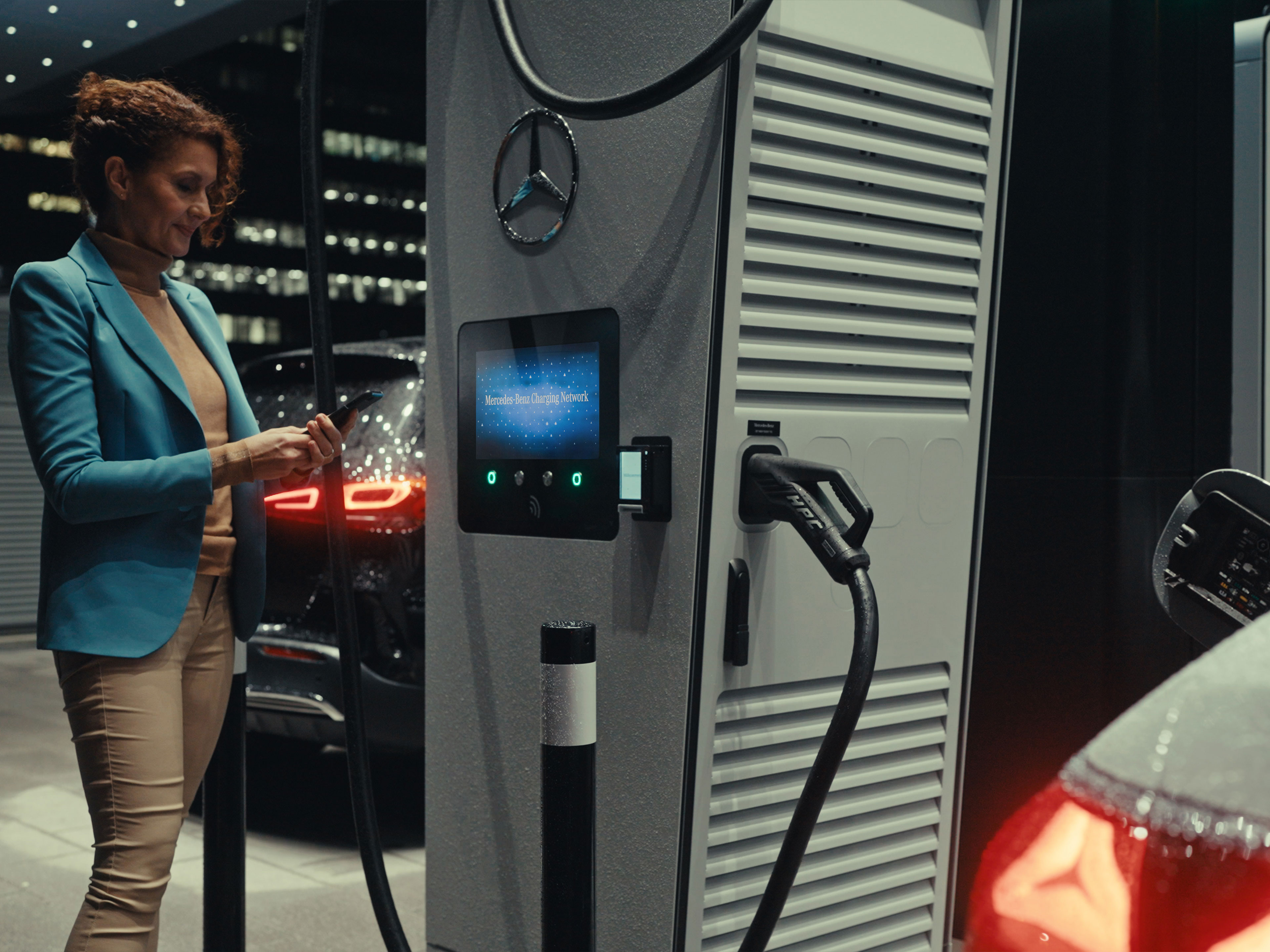 Application Mercedes-Benz | Recharge en déplacement | Mercedes-Benz Une femme tenant un smartphone à la main, debout à côté d’une station de recharge.
