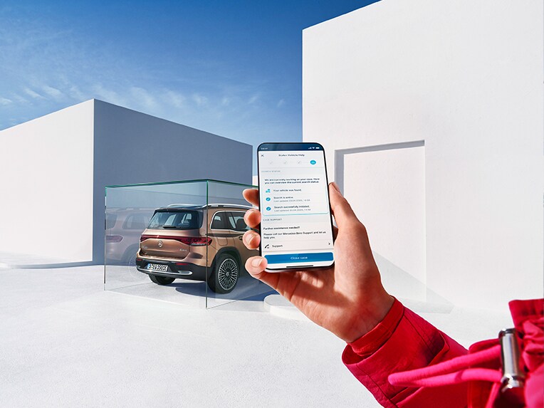 Une personne tenant un téléphone portable dans le téléphone portable et utilisant l'application Mercedes. Un EQB stationne en arrière-plan.