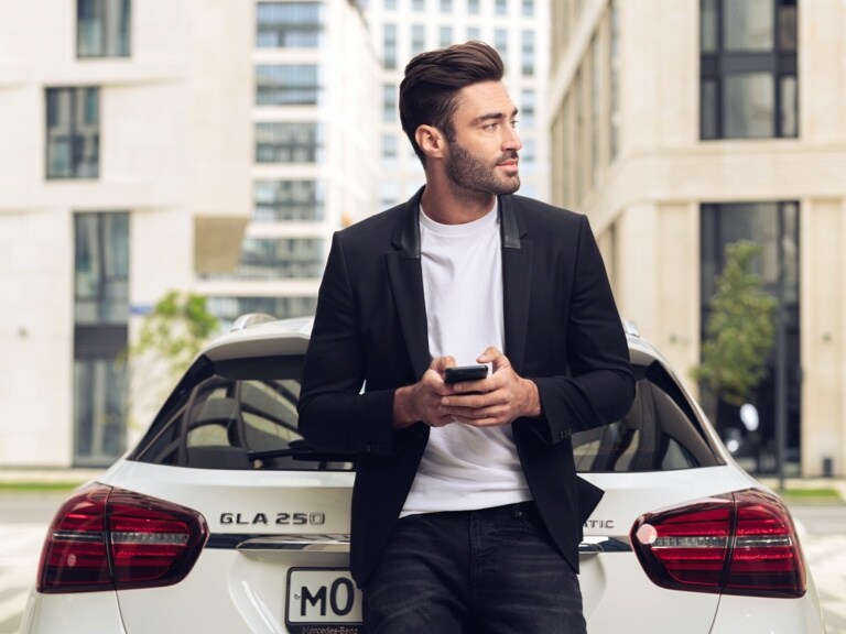 Un homme est accoudé à sa Mercedes-Benz GLA et s’informe sur la météo de son lieu d’arrivée via l’application Mercedes me sur son smartphone et les offres digitales sur le thème de la navigation.