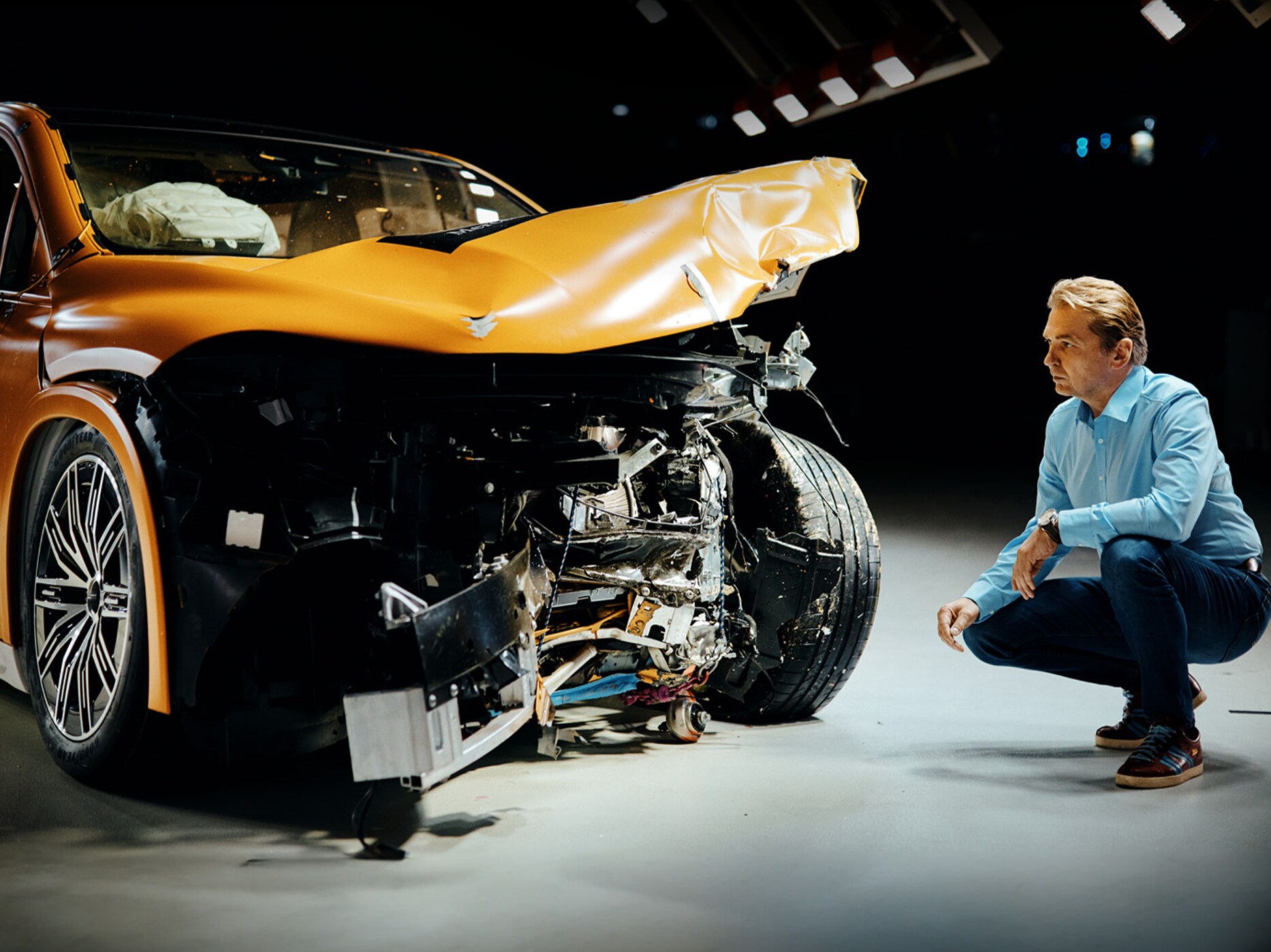 Un expert en essais de collision examine une voiture Mercedes-Benz accidentée à l'avant en s'accroupissant à côté d'elle.