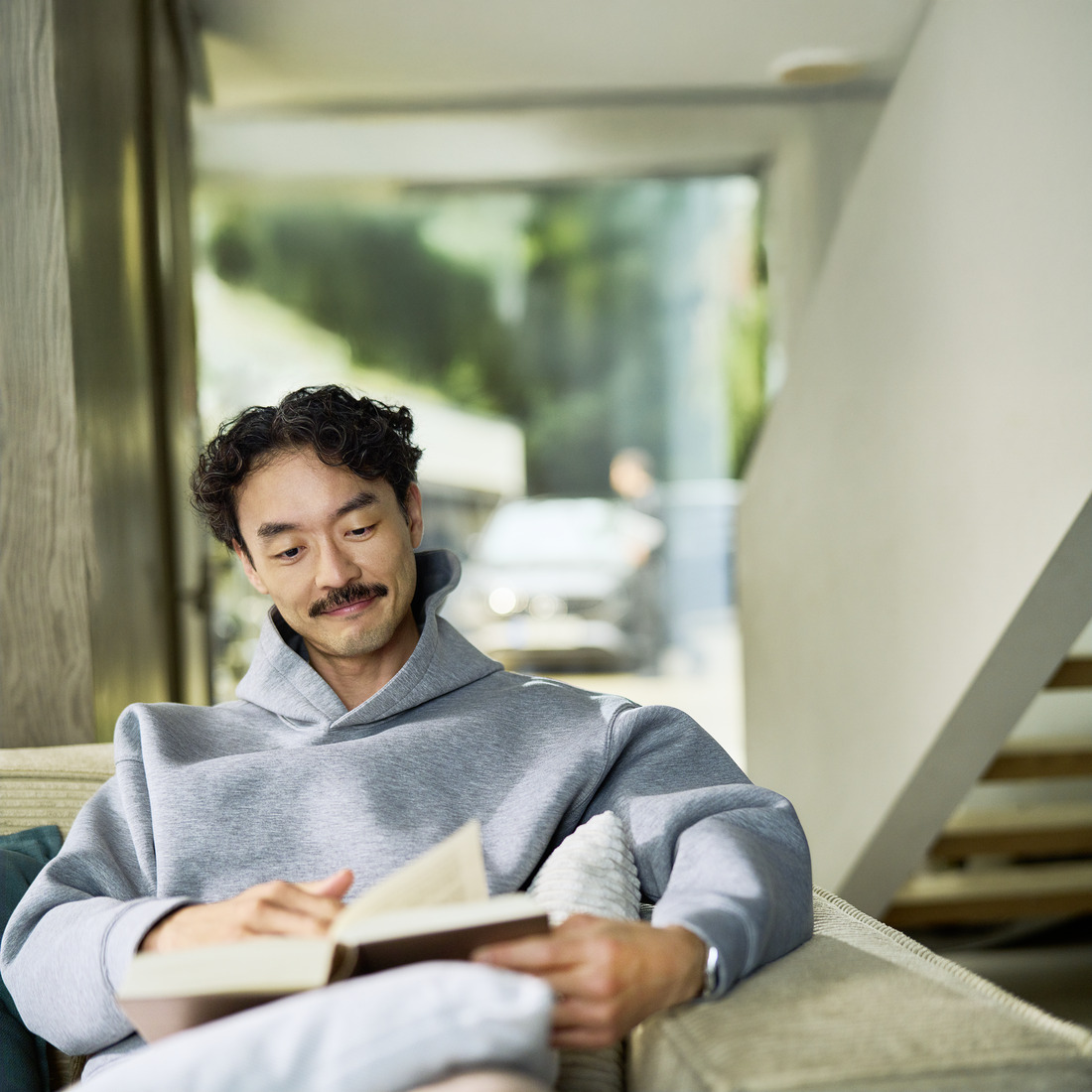 Client détendu bénéficiant du Mobile Service à domicile Client assis à l'intérieur en train de lire un livre. Derrière lui, on aperçoit à travers une fenêtre une voiture en cours d'entretien par un spécialiste.