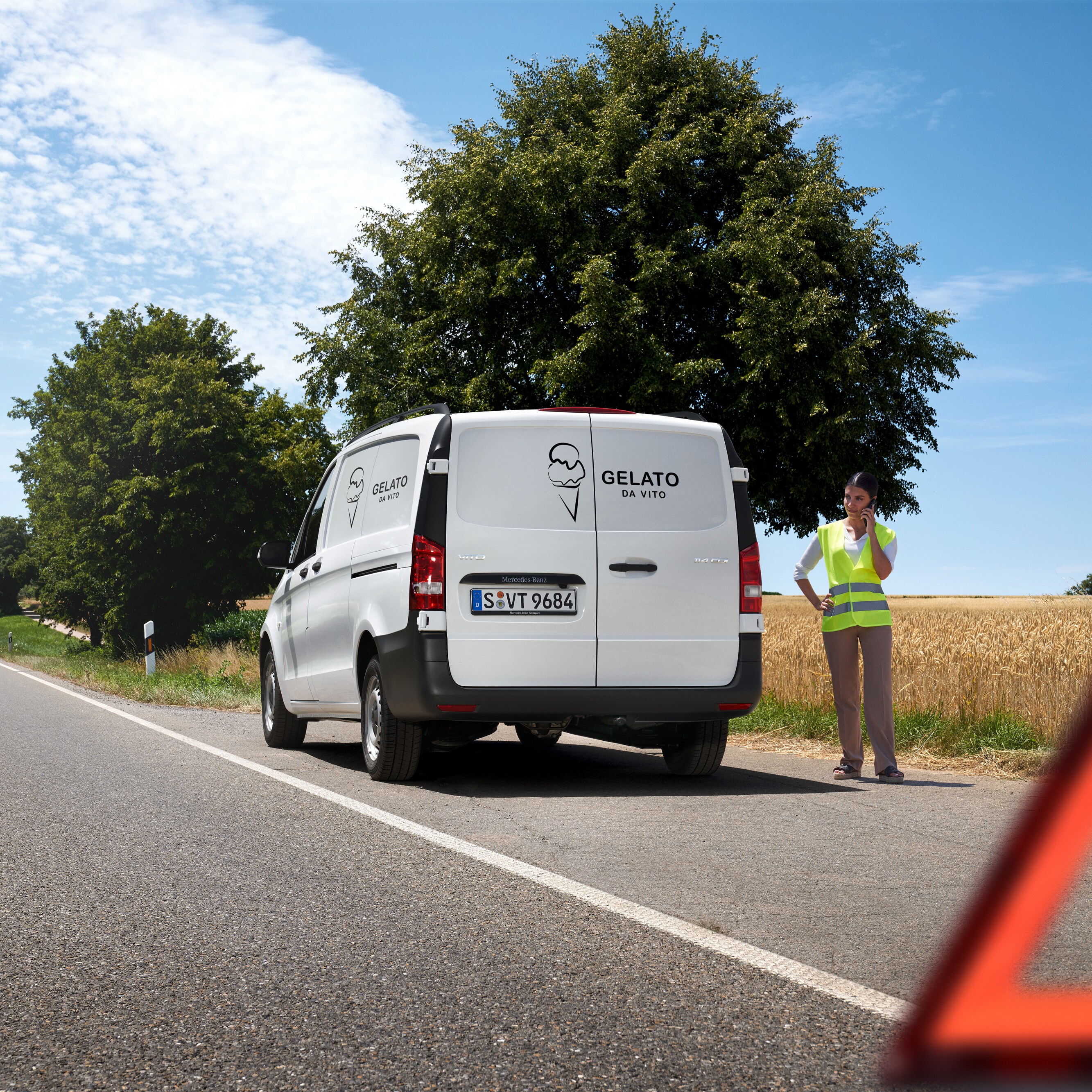 Een vrouw belt de Mercedes-Benz Service24h-hotline, terwijl een Mercedes-Benz Sprinter langs een landweg stilstaat.