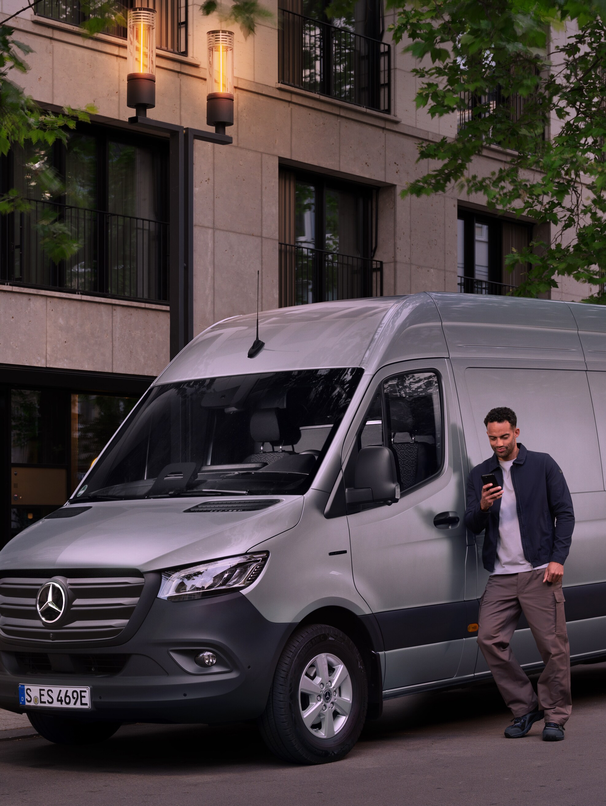 Man leunt tegen een zilveren Mercedes-Benz Sprinter Gesloten Bestelwagen en gebruikt zijn smartphone – snelle hulp van Mercedes-Benz Klantenzorg.