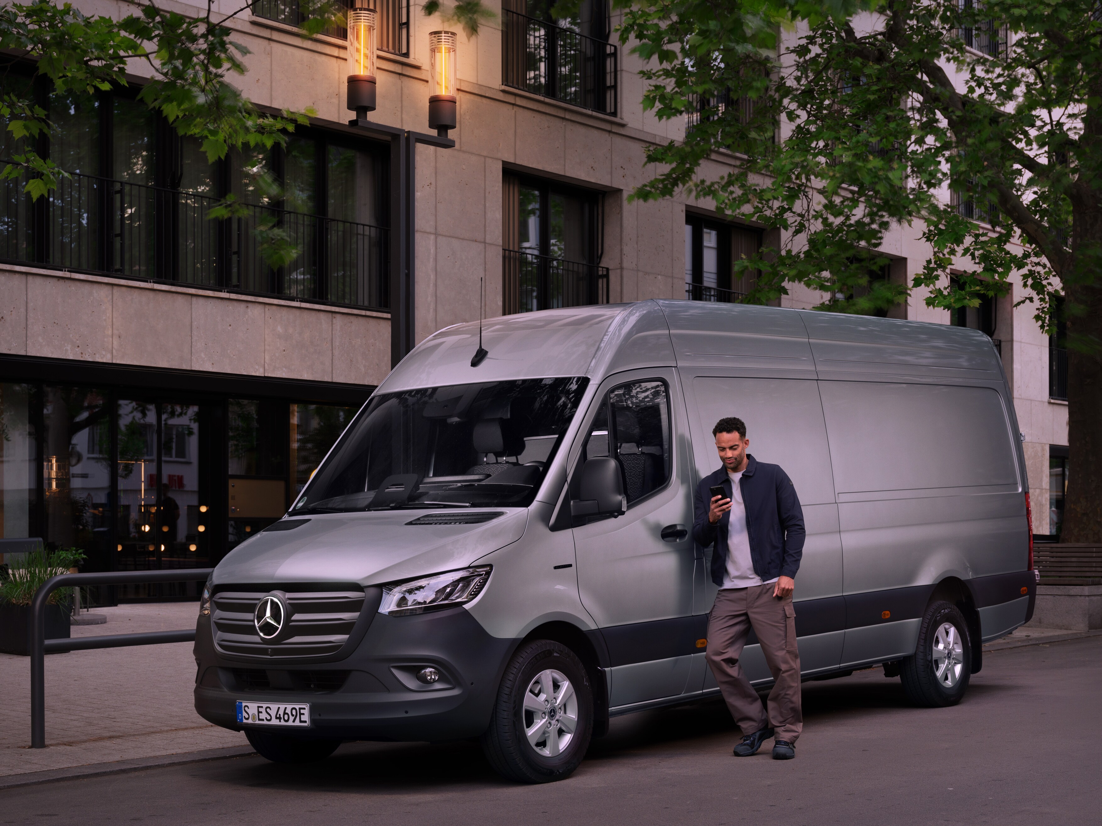 Man leunt tegen een zilveren Mercedes-Benz Sprinter Gesloten Bestelwagen en gebruikt zijn smartphone – snelle hulp van Mercedes-Benz Klantenzorg.