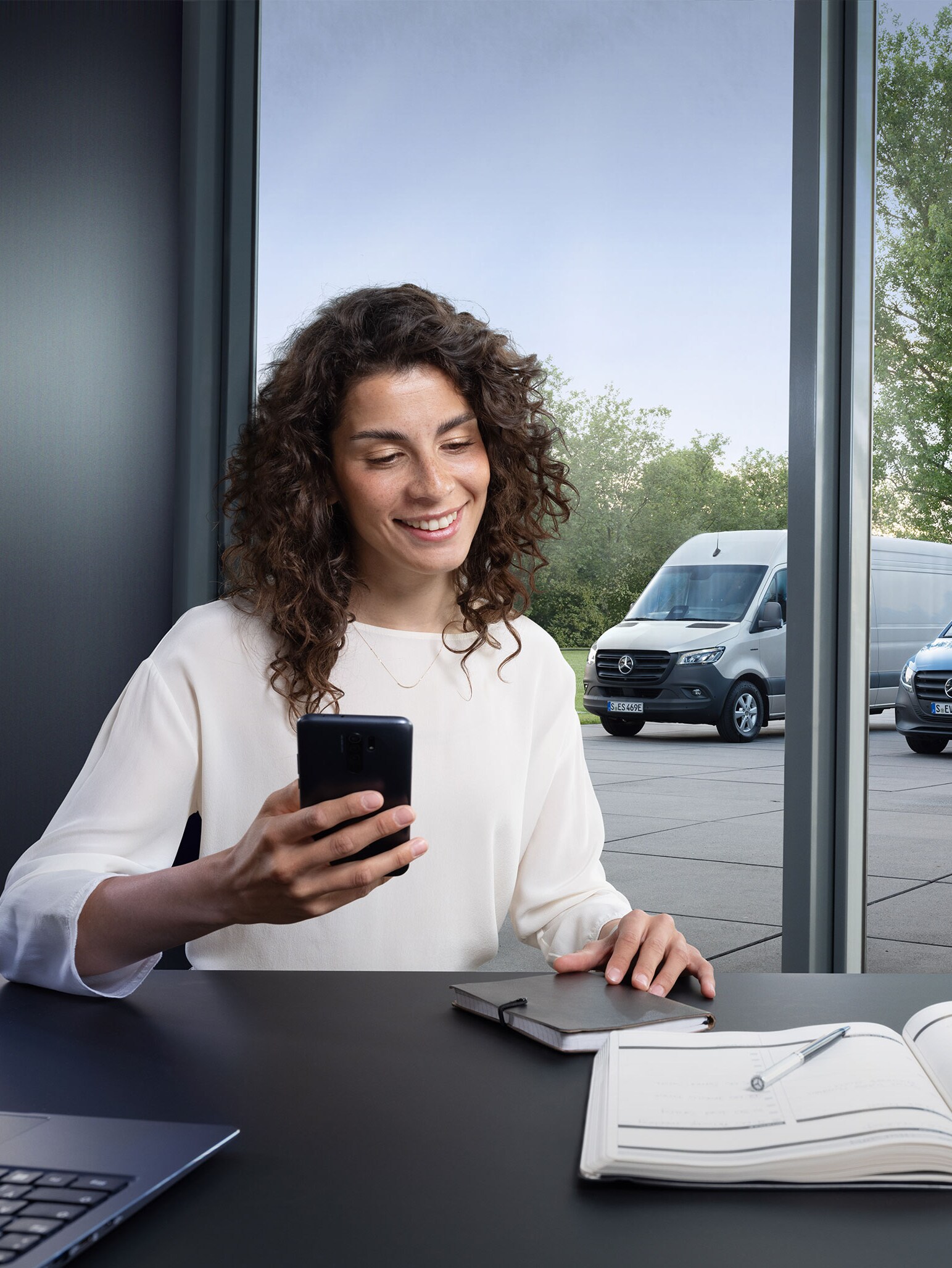 Vrouw met smartphone aan een bureau, met meerdere Mercedes-Benz Bedrijfswagens op de achtergrond – deskundige ondersteuning in Business Care.