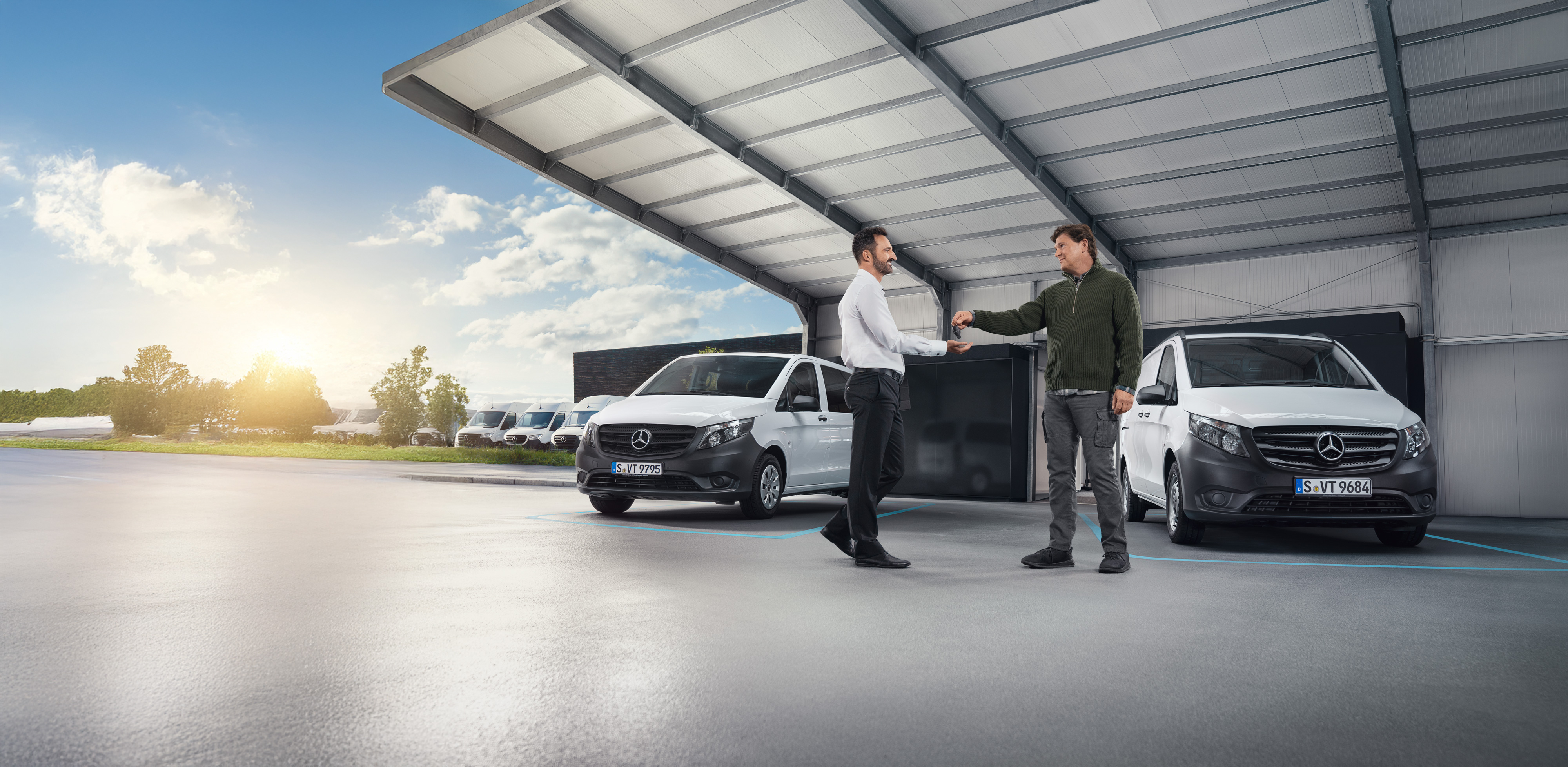 Mercedes-Benz Bedrijfswagen | Vans Care | Individuele Ondersteuning Twee mannen in gesprek voor een reeks witte Mercedes-Benz Bedrijfswagens in een hal.