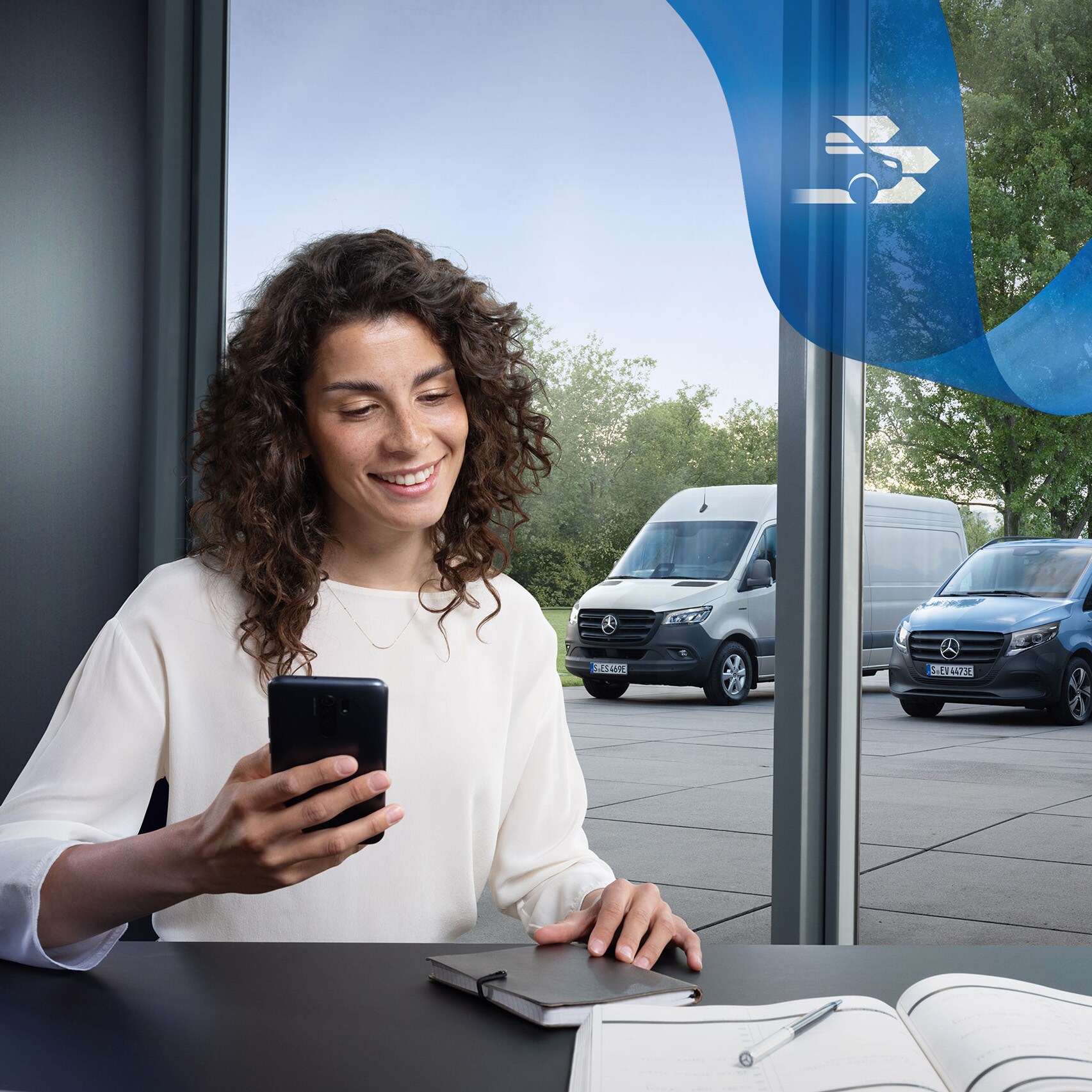 Vrouw zittend aan een bureau met een smartphone en een reeks Mercedes-Benz Bedrfijswagens op de achtergrond.