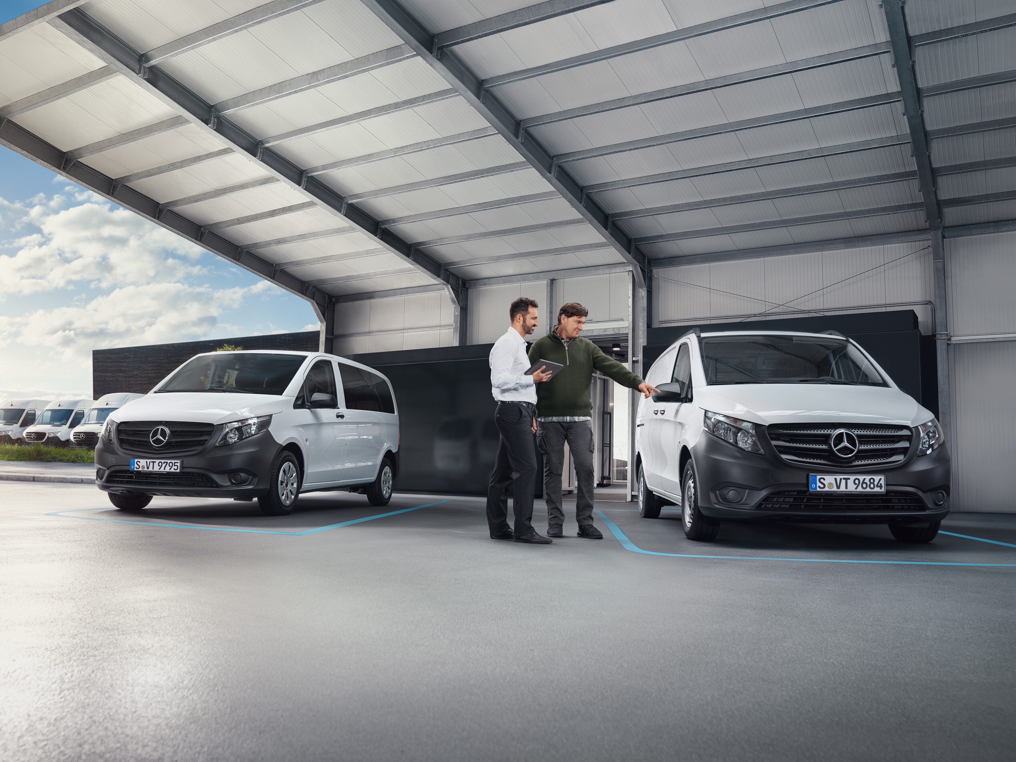 Twee personen in een werkplaats in gesprek, staand voor twee witte Mercedes-Benz bedrijfswagens.