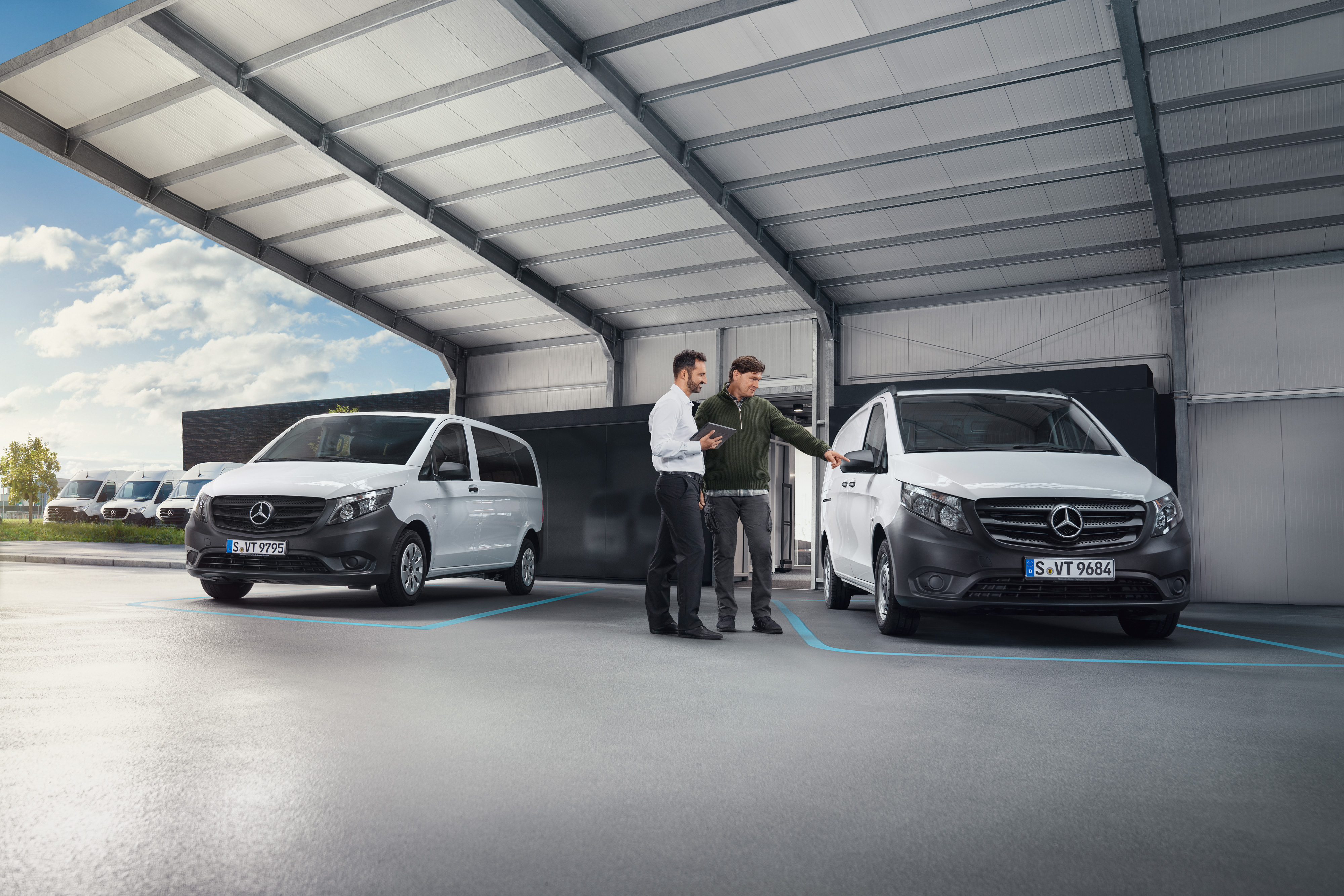 Mercedes-Benz | Comfort | Digitale klantenreceptie Twee personen in een werkplaats in gesprek, staand voor twee witte Mercedes-Benz bedrijfswagens.
