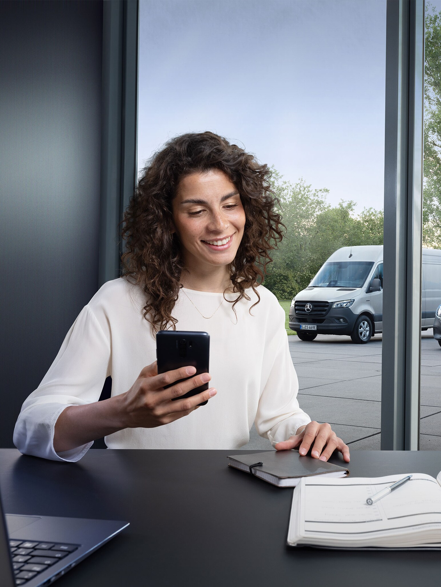 Vrouw boekt een service via smartphone met een reeks Mercedes-Benz bedrijfswagens op de achtergrond.