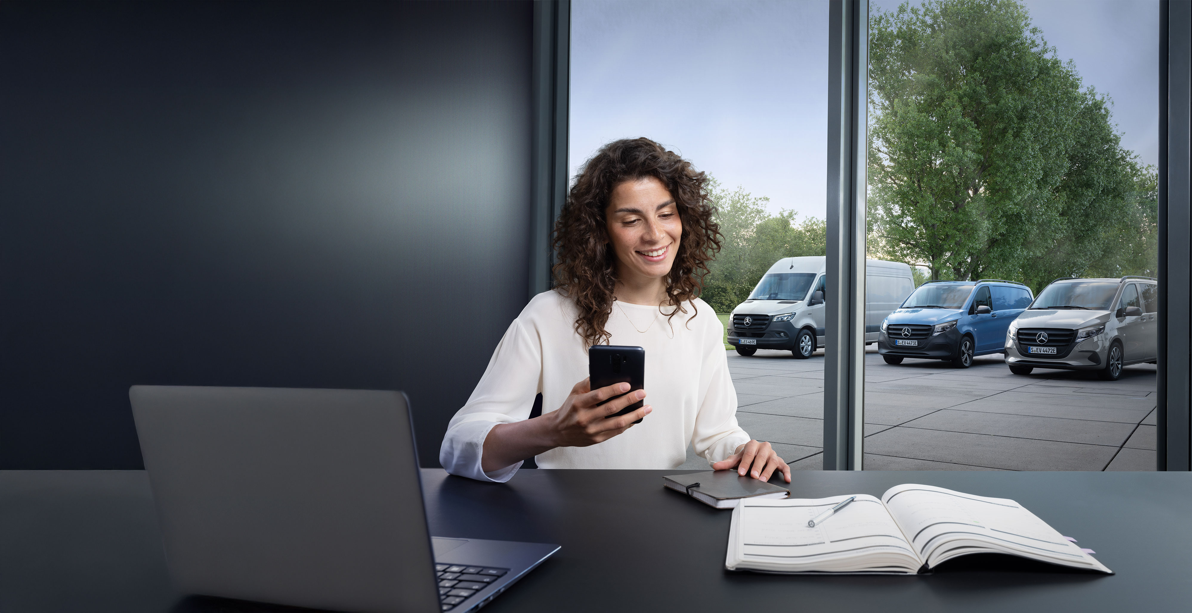 Mercedes-Benz | Comfort | Online Afspraak Reserveren Vrouw boekt een service via smartphone met een reeks Mercedes-Benz bedrijfswagens op de achtergrond.