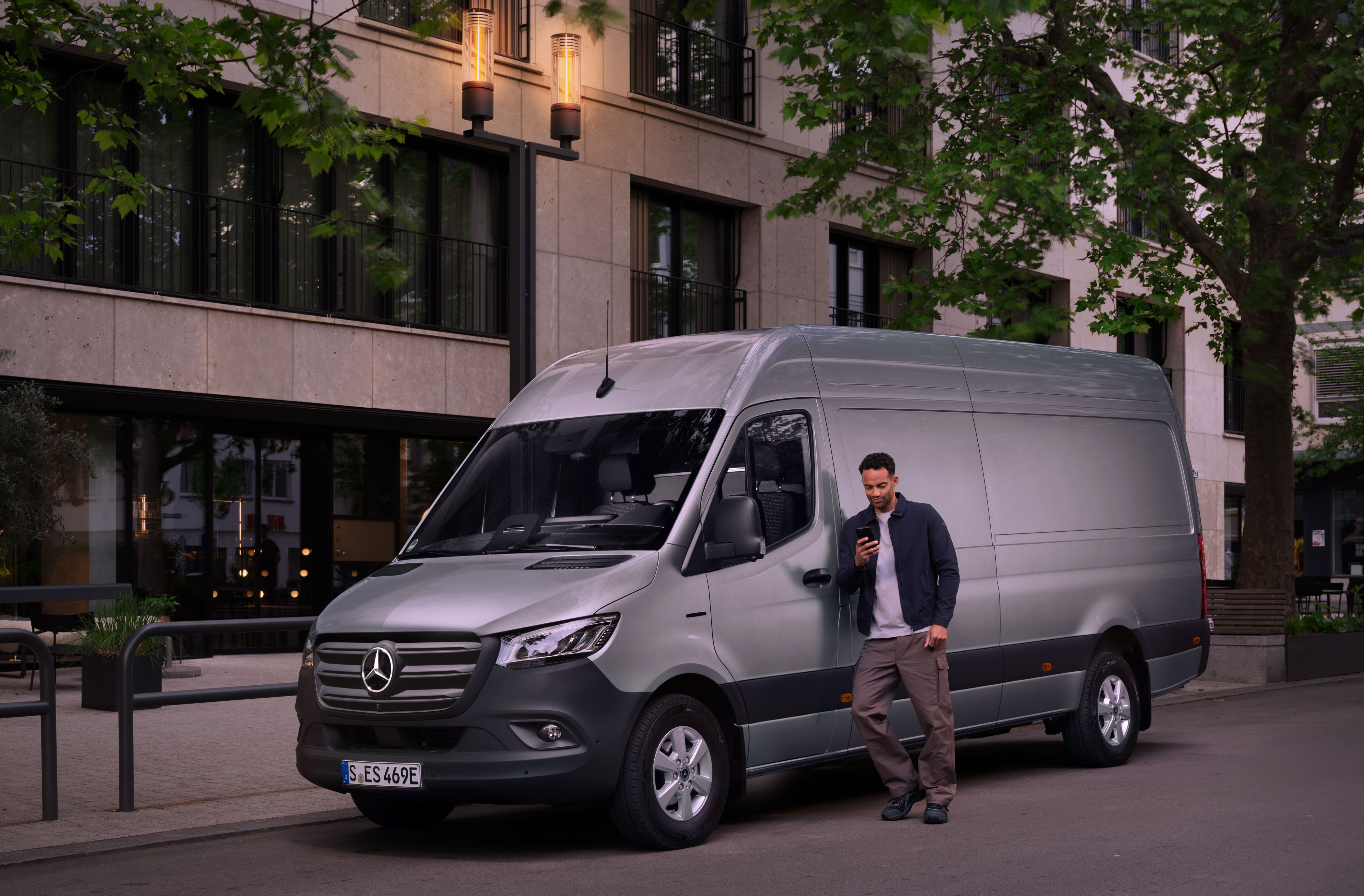 Mercedes-Benz Bedrijfswagen | Reparatie | Digitaal Schaderapport Man leunt tegen zilveren Mercedes-Benz Sprinter en vult digitaal schaderapport in op zijn smartphone.