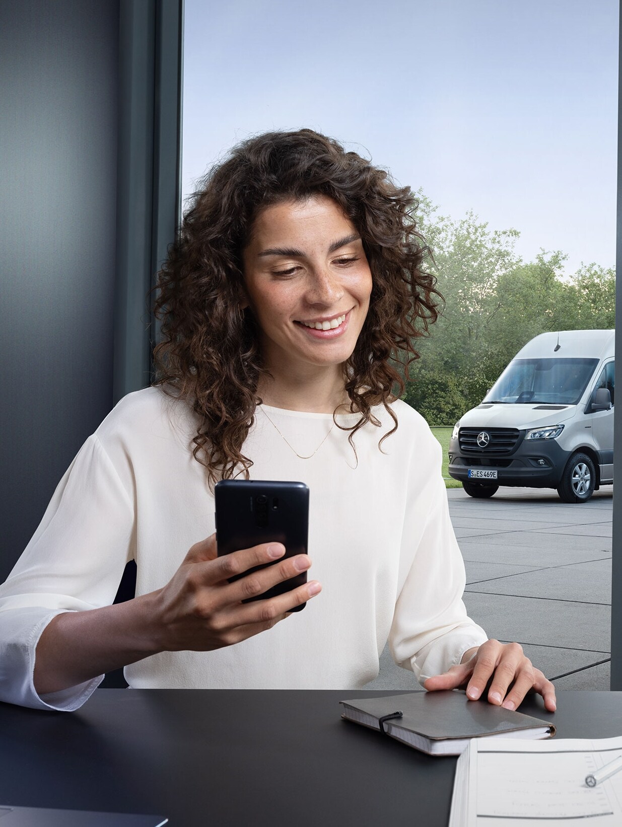 Vrouw die met een smartphone het wagenparkbeheer van meerdere Mercedes-Benz bedrijfswagens bedient bij het kantoorraam.